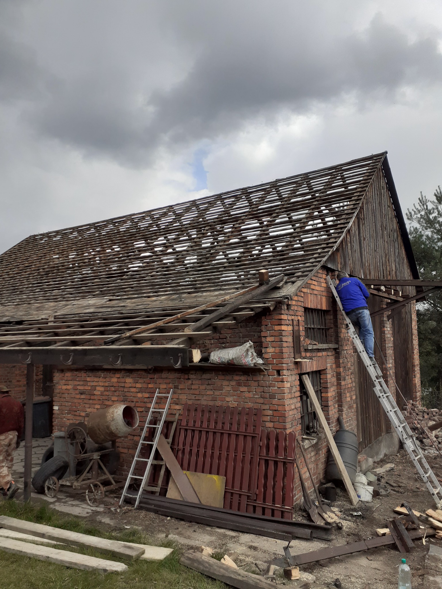 Ceglany budynek z częściowo zdjętym dachem, pracownik na drabinie w niebieskiej bluzie, stara betoniarka i porozrzucane drewniane elementy na trawie.