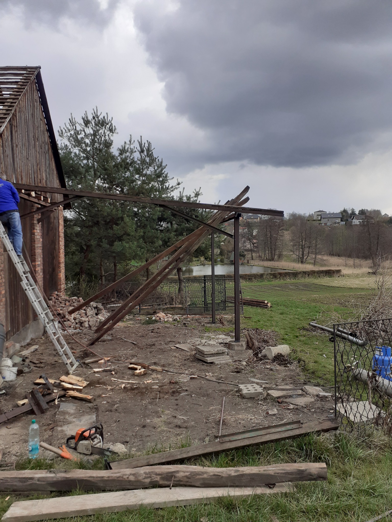 Demontaż drewnianej konstrukcji budynku gospodarczego na tle pochmurnego nieba, z widocznymi elementami rozbiórki, takimi jak porozrzucane cegły i drewno, oraz narzędzia: piła spalinowa i drabina.