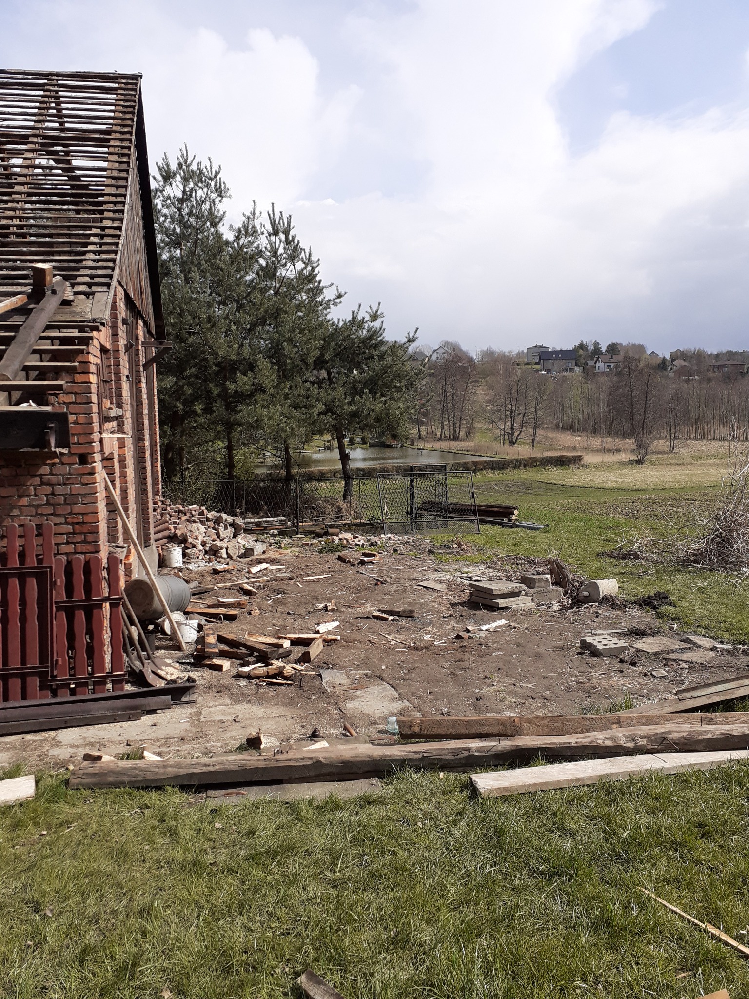 Częściowo rozebrany, ceglany budynek z odsłoniętą konstrukcją dachu, gruz i drewniane elementy leżące na ziemi, widok na okoliczny krajobraz z drzewami i zbiornikiem wodnym w tle.