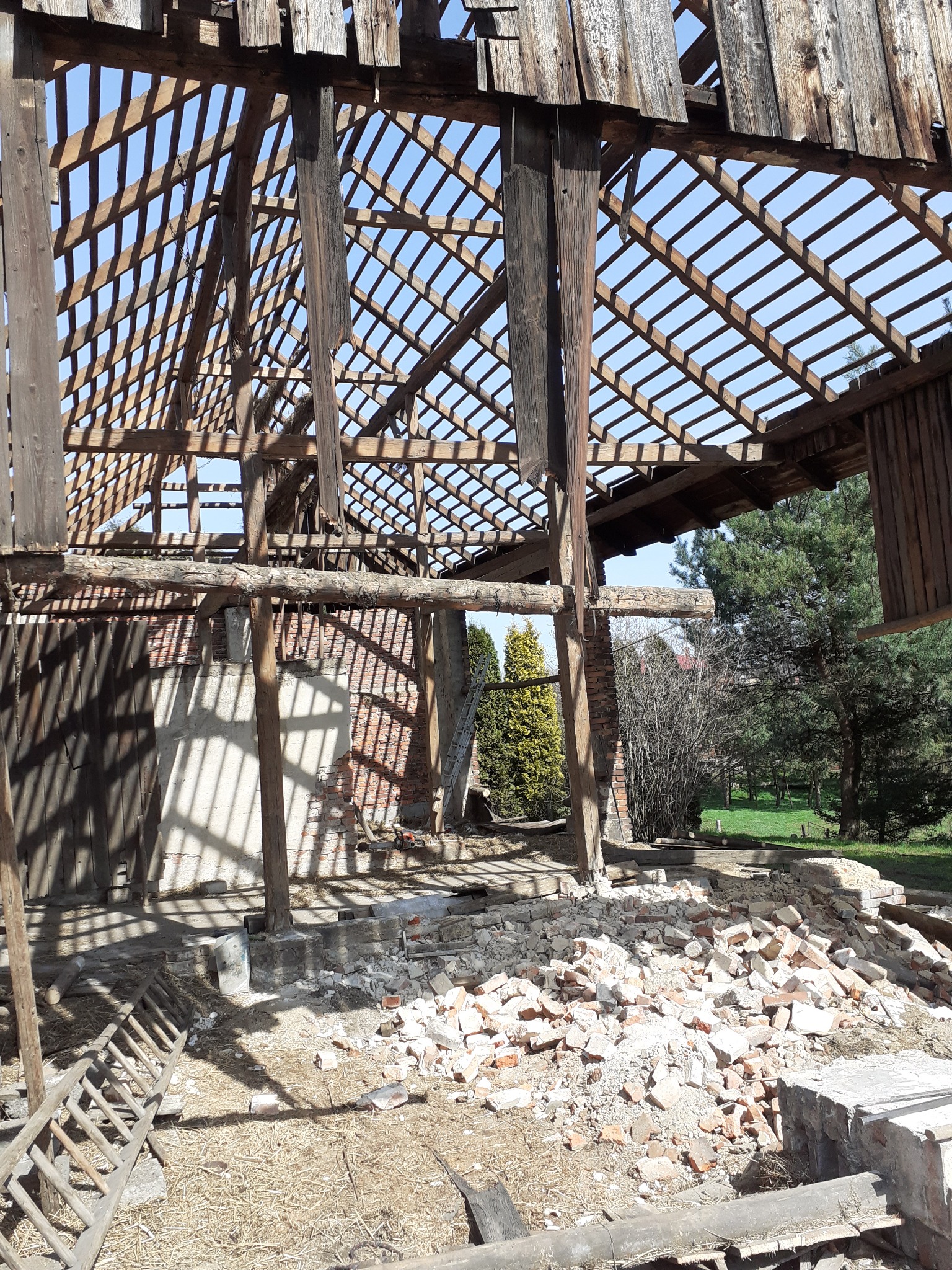 Zdemontowana konstrukcja drewniana dachu starego budynku z fragmentami muru i gruzem ceglanym, widok z dołu na niebo przez kratownicę belek.