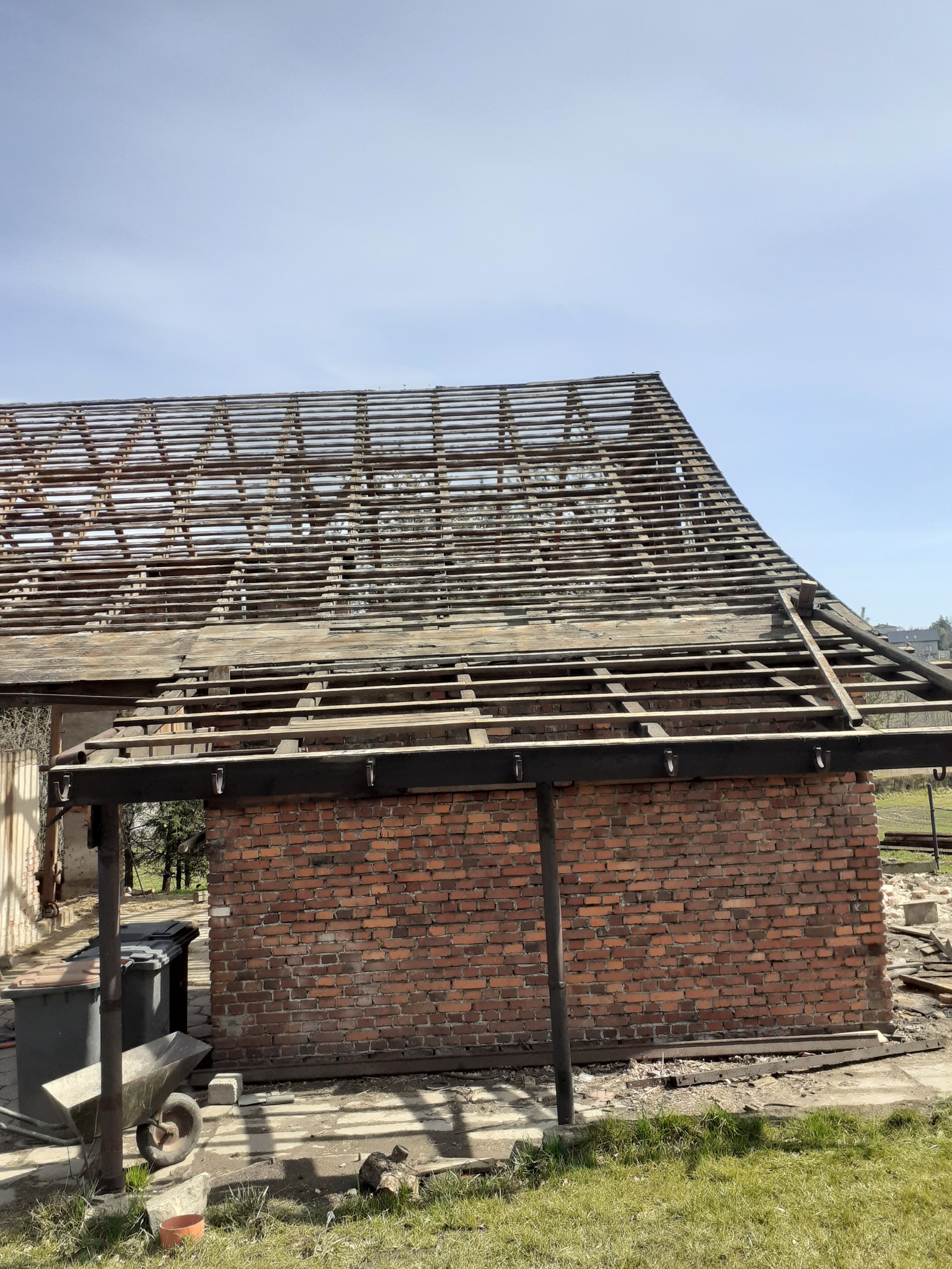 Ceglany budynek bez dachu, z widoczną konstrukcją więźby dachowej, w tle zieleń i błękitne niebo.