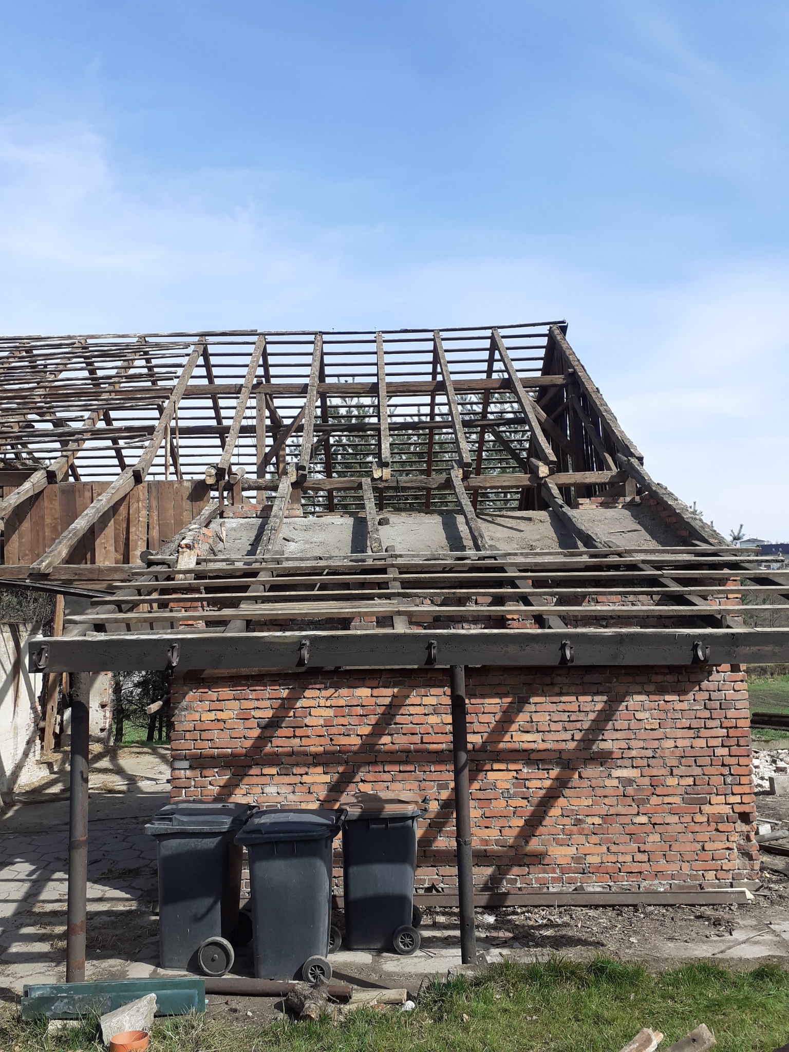 Ceglany budynek w trakcie rozbiórki, widoczne fragmenty dachu i ścian, trzy czarne pojemniki na śmieci na pierwszym planie, błękitne niebo w tle.