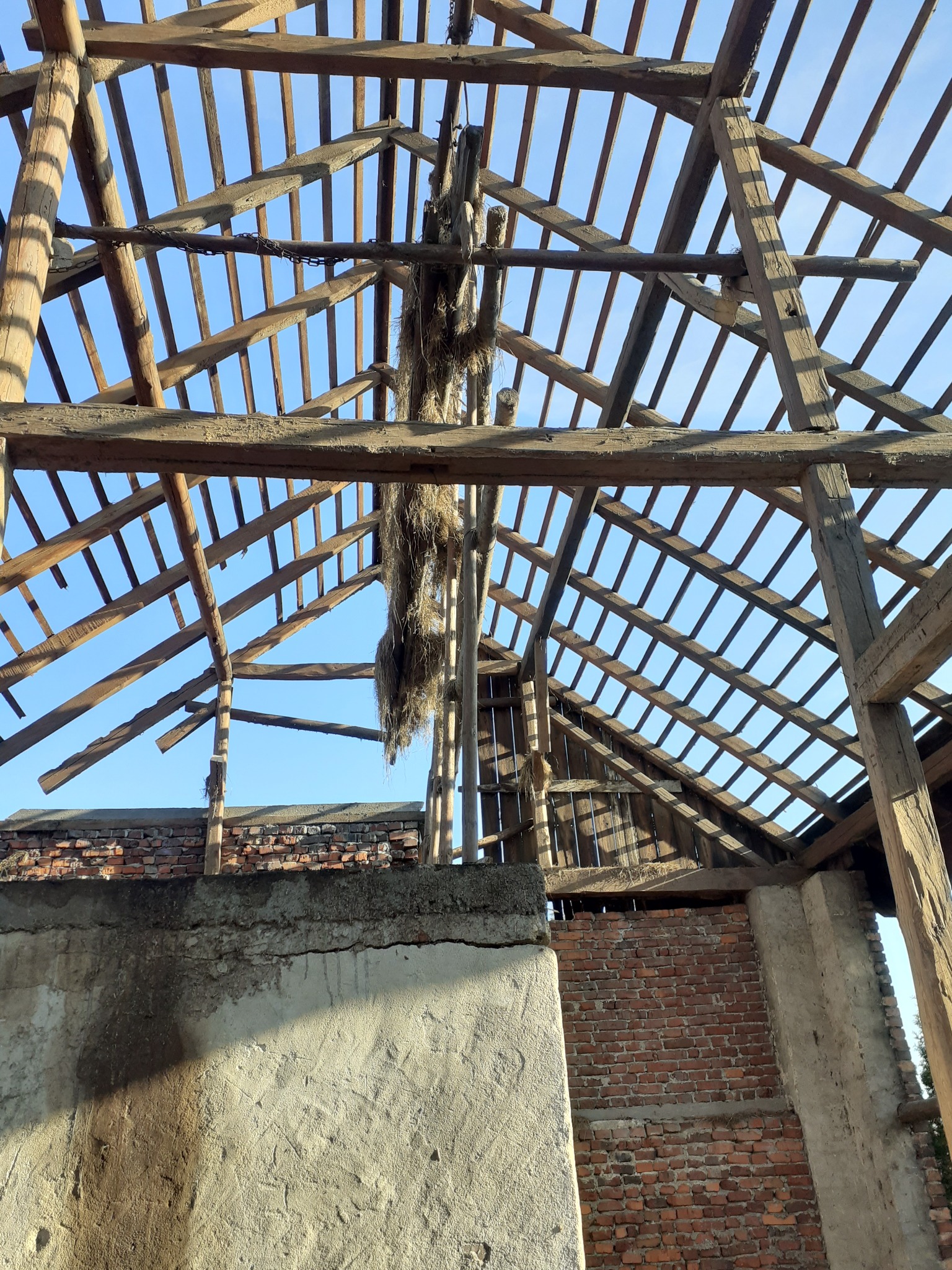 Zdjęcie z dołu na drewnianą konstrukcję dachu starego budynku w trakcie rozbiórki, z widocznymi elementami muru i niebem w tle.
