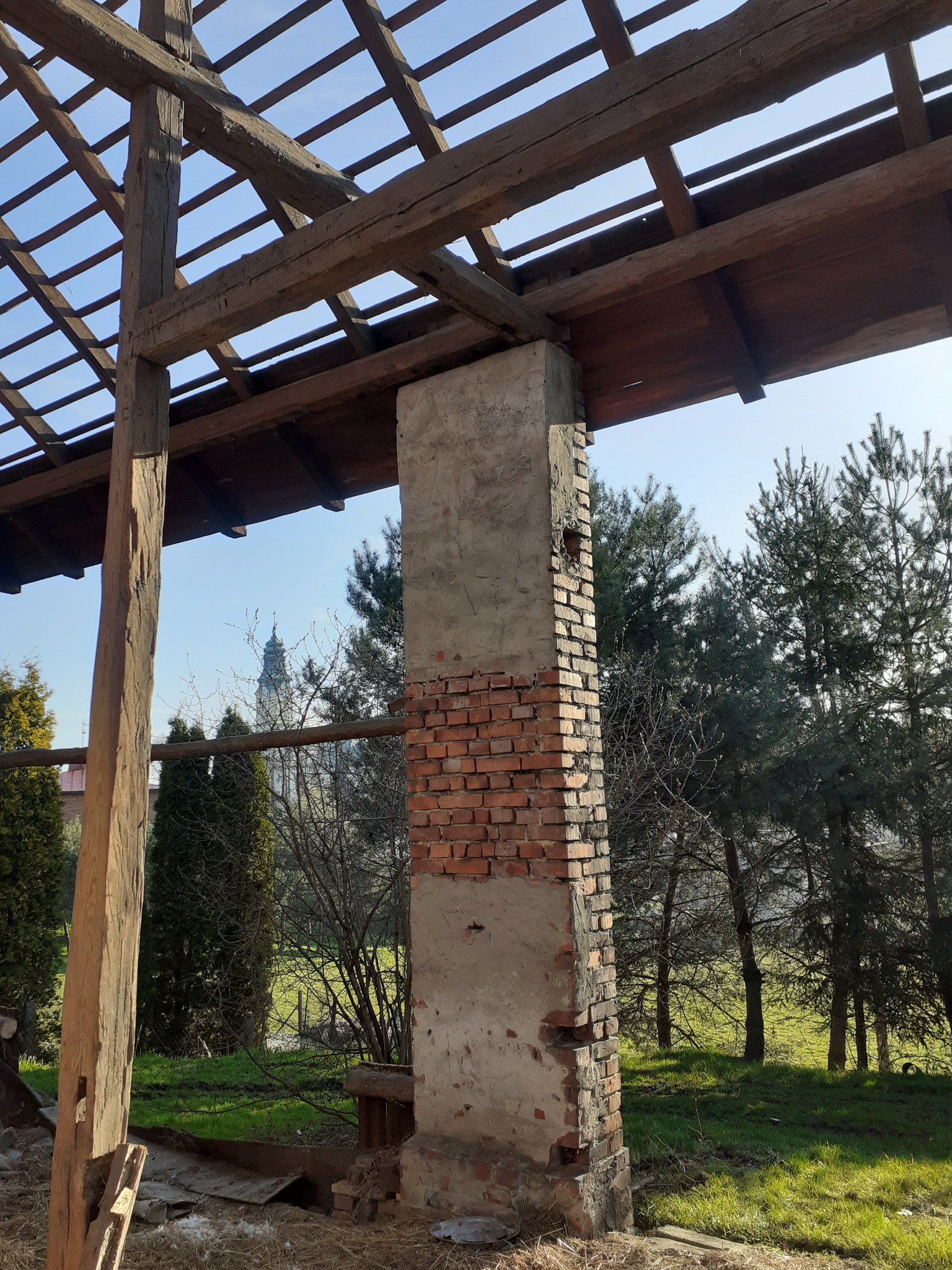 Częściowo rozebrany budynek z drewnianą konstrukcją dachu, fragment murowanej kolumny z cegły i betonu, widok na oddalony kościół przez otwór w ścianie, drzewa w tle.