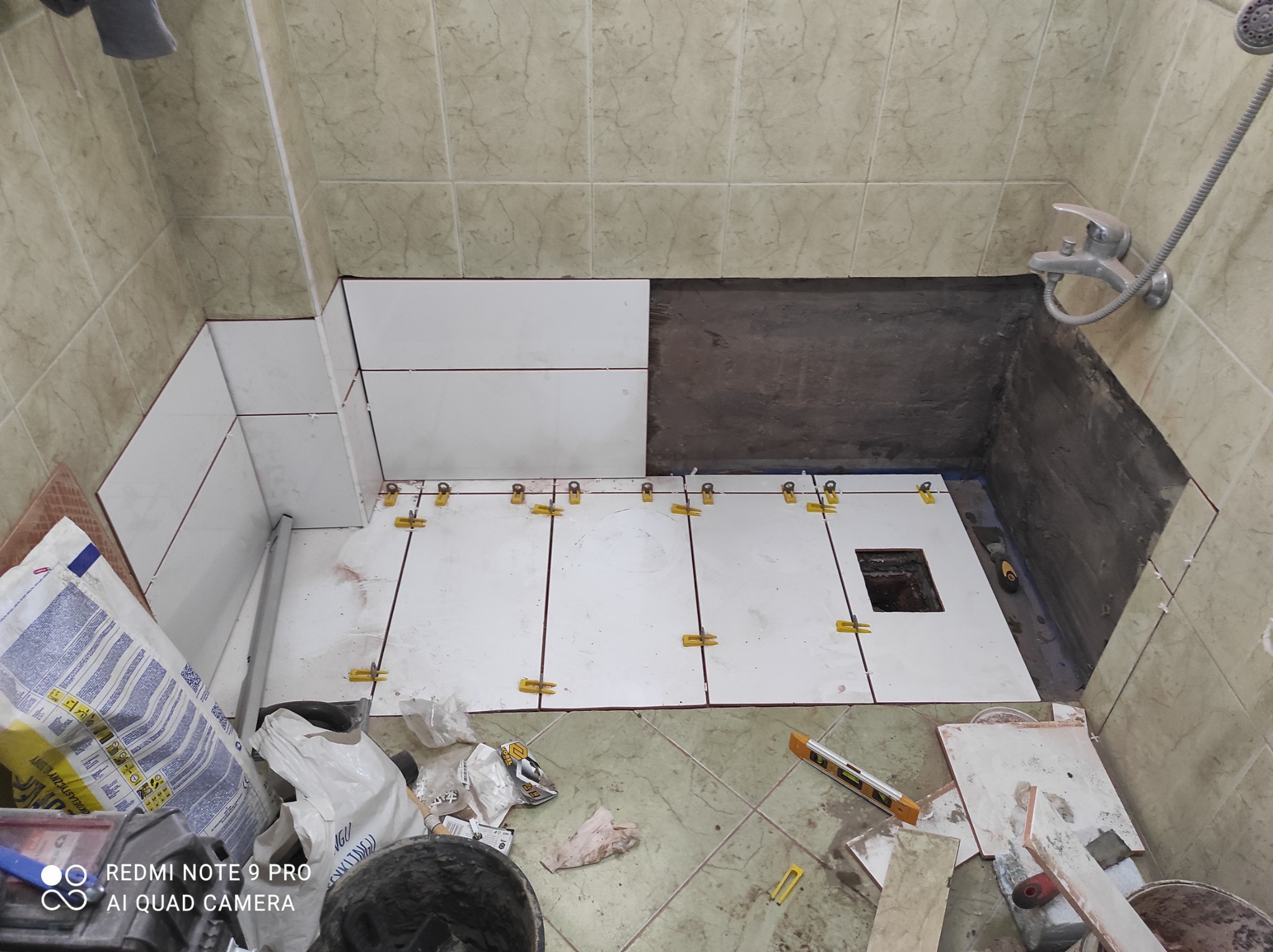 Układanie białych płytek w łazience, widoczne żółte klipsy poziomujące, częściowo wykończona ściana prysznica, worki z zaprawą i poziomica na podłodze.