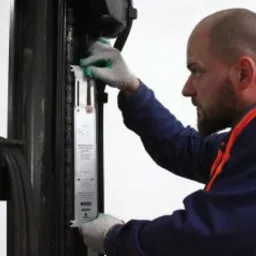 Mężczyzna w roboczych rękawicach i kamizelce odblaskowej sprawdza stan techniczny wózka widłowego, koncentrując się na elementach łańcucha i tabliczce informacyjnej.