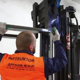 Instruktor w pomarańczowej kamizelce z napisem 'AWANS B.H.P' demonstruje spawanie elementu wózka widłowego, nosi rękawice ochronne, widoczne iskry spawania.