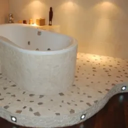 Nowoczesna łazienka z wanną obudowaną mozaiką, podłoga z płytek w odcieniach beżu i brązu z wbudowanymi punktowymi lampkami, jasne ściany i dekoracje na półce.