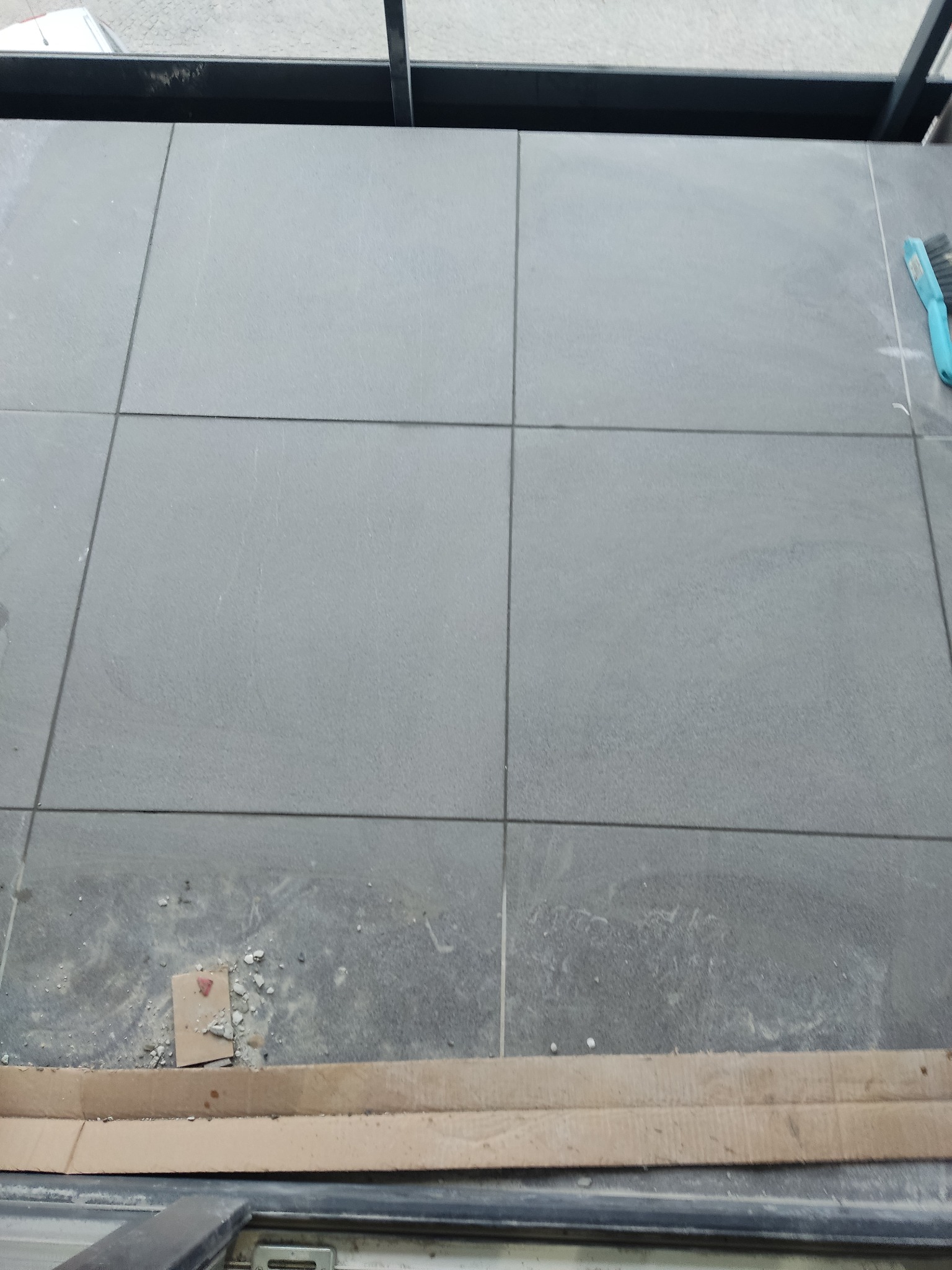 Szare płytki podłogowe ułożone na balkonie, widoczna szczotka do fugowania i resztki zaprawy.