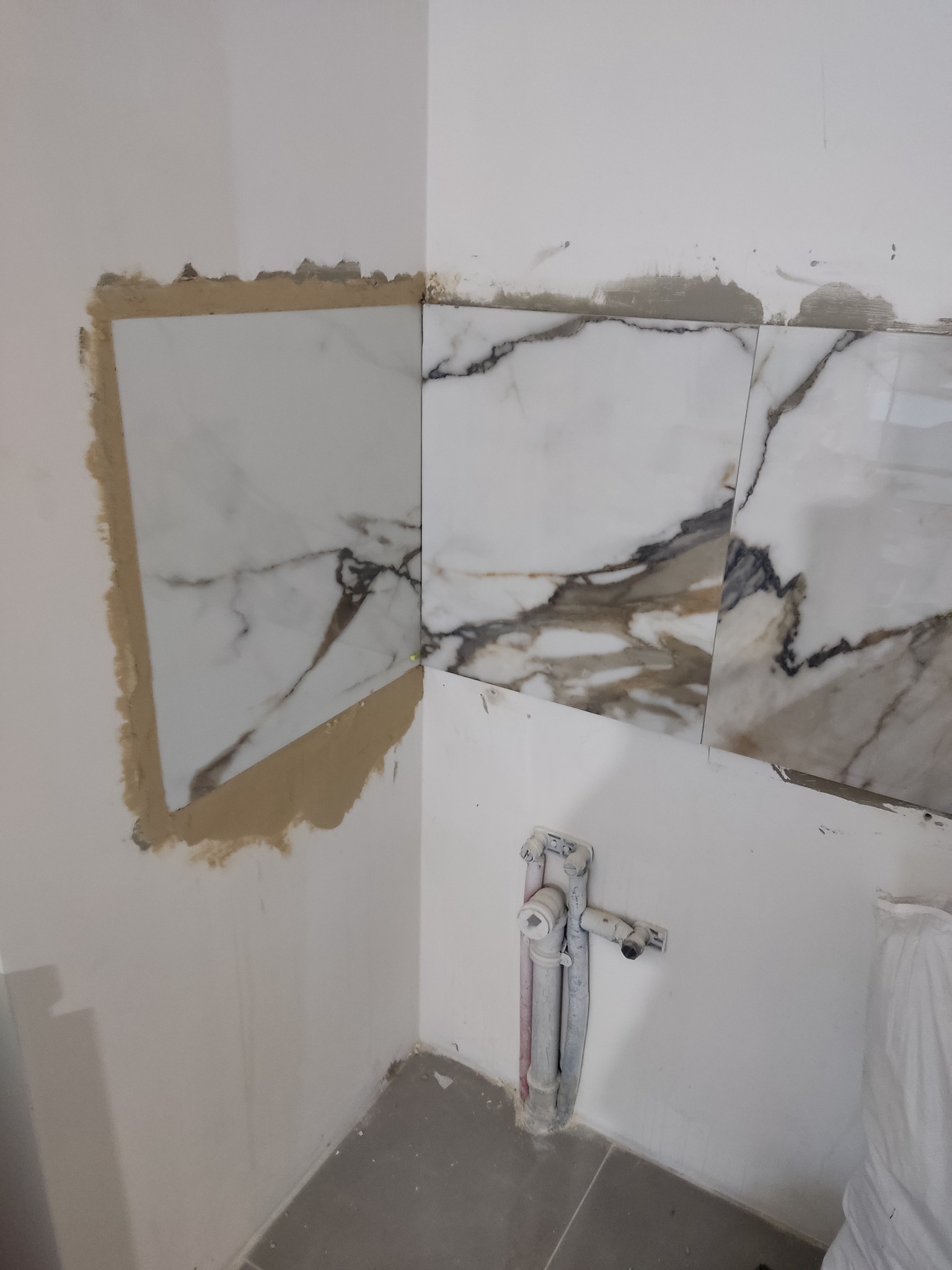 Fragment ściany w trakcie układania płytek imitujących marmur, widoczny klej i rury instalacyjne.