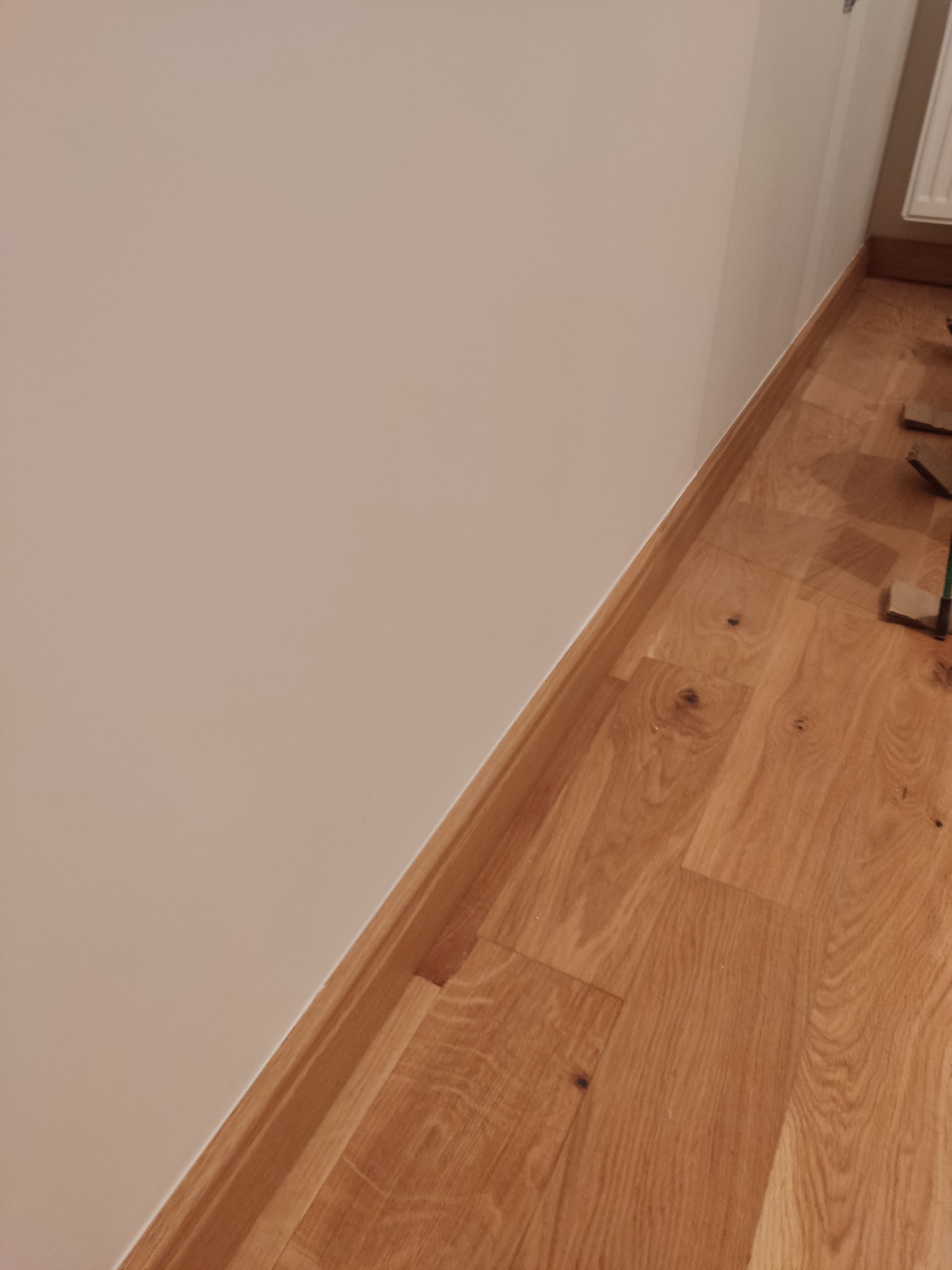 Jasne panele podłogowe z drewnianą listwą przypodłogową przy beżowej ścianie, widoczne fragmenty narzędzi w prawym górnym rogu.