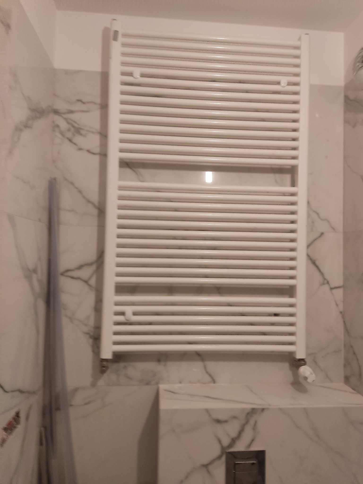 Biały grzejnik drabinkowy zamontowany na ścianie wyłożonej marmurowymi płytkami w łazience, widoczne elementy instalacji i fragment kabiny prysznicowej.