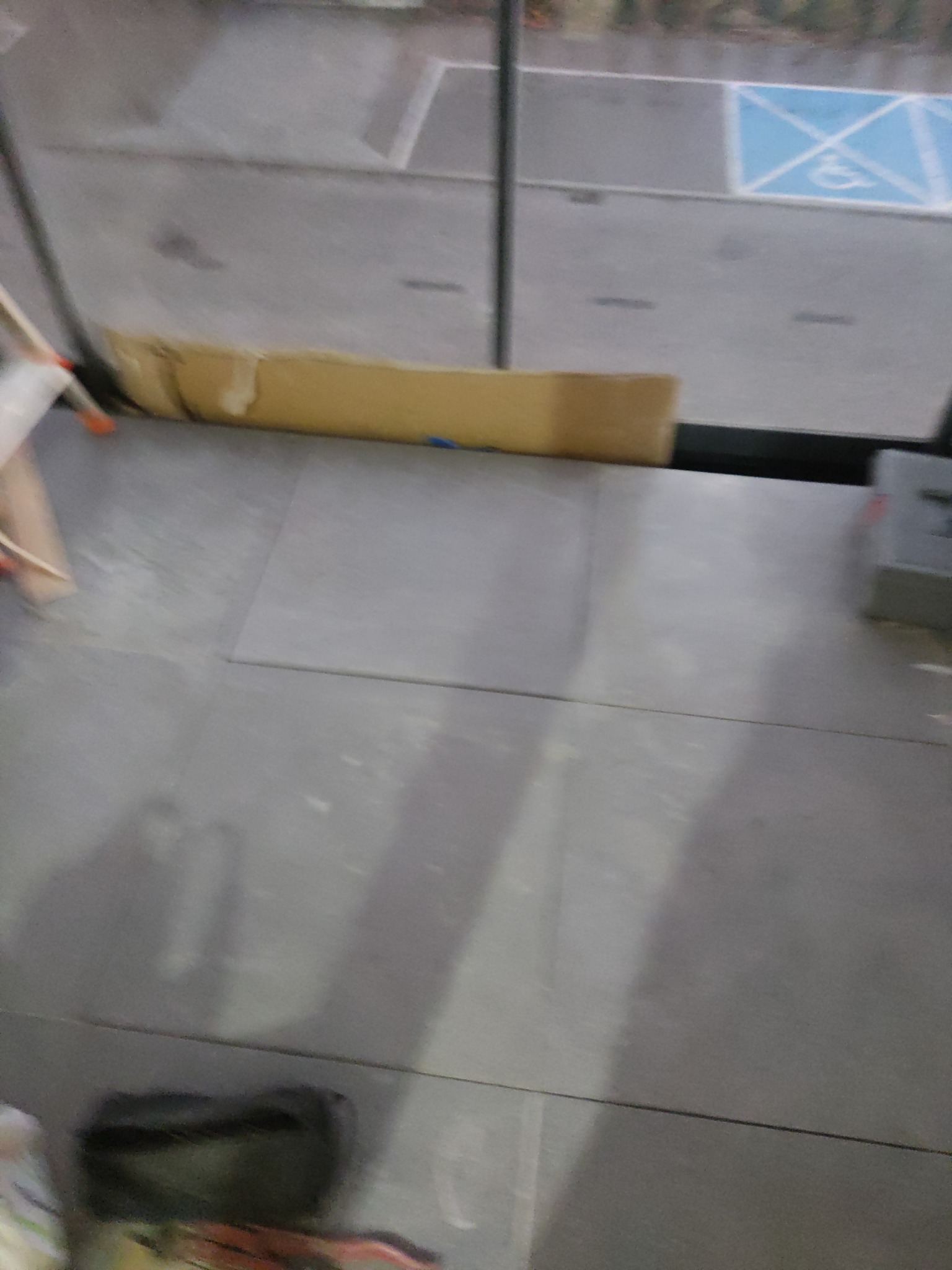 Szare płytki podłogowe, częściowo ułożone, z widocznym kartonem ochronnym przy oknie i fragmentem parkingu z symbolem osoby niepełnosprawnej w tle.