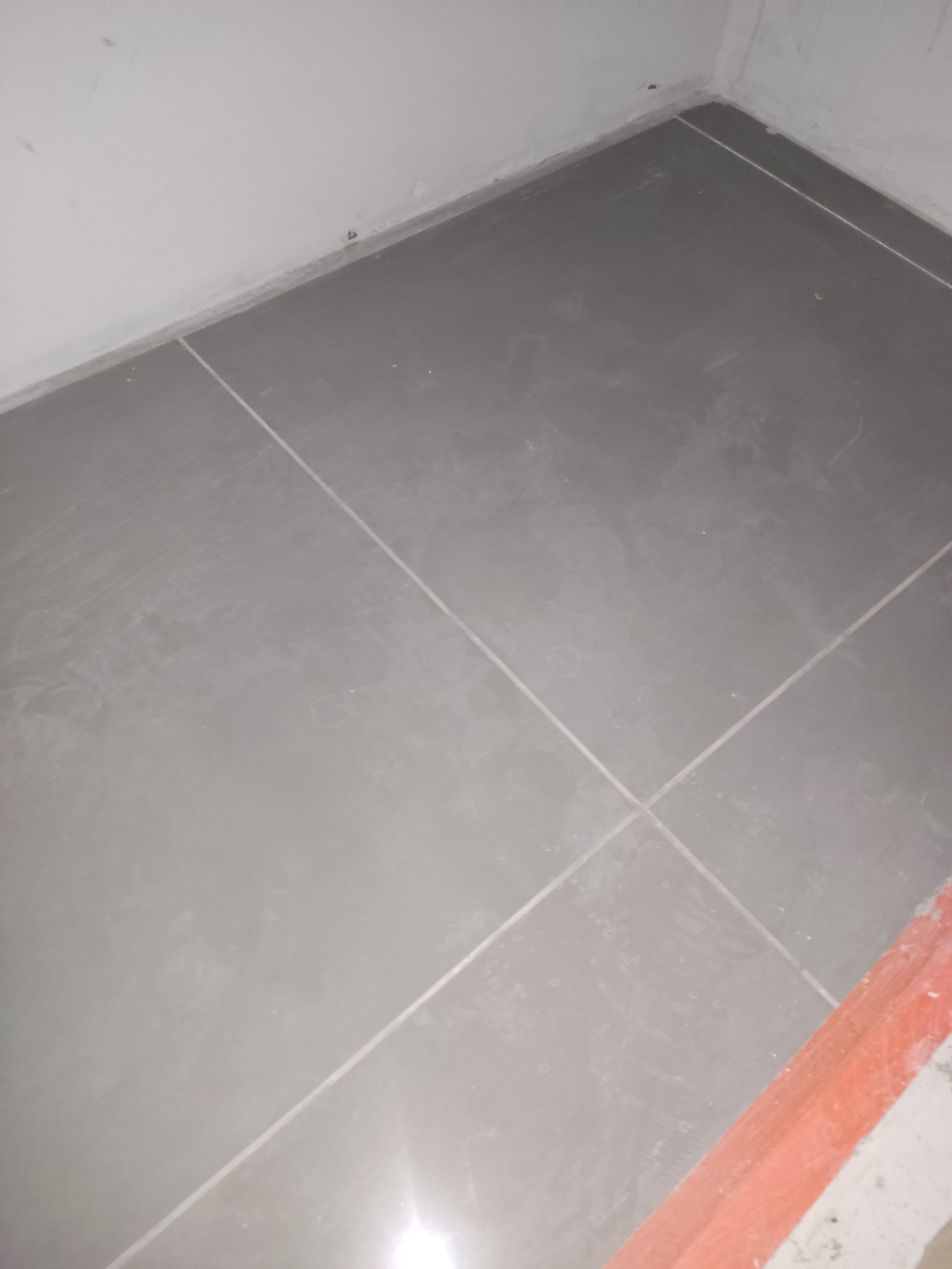 Świeżo ułożone, szare płytki podłogowe z jasną fugą w rogu pomieszczenia, widoczny fragment pomarańczowego profilu wykończeniowego.