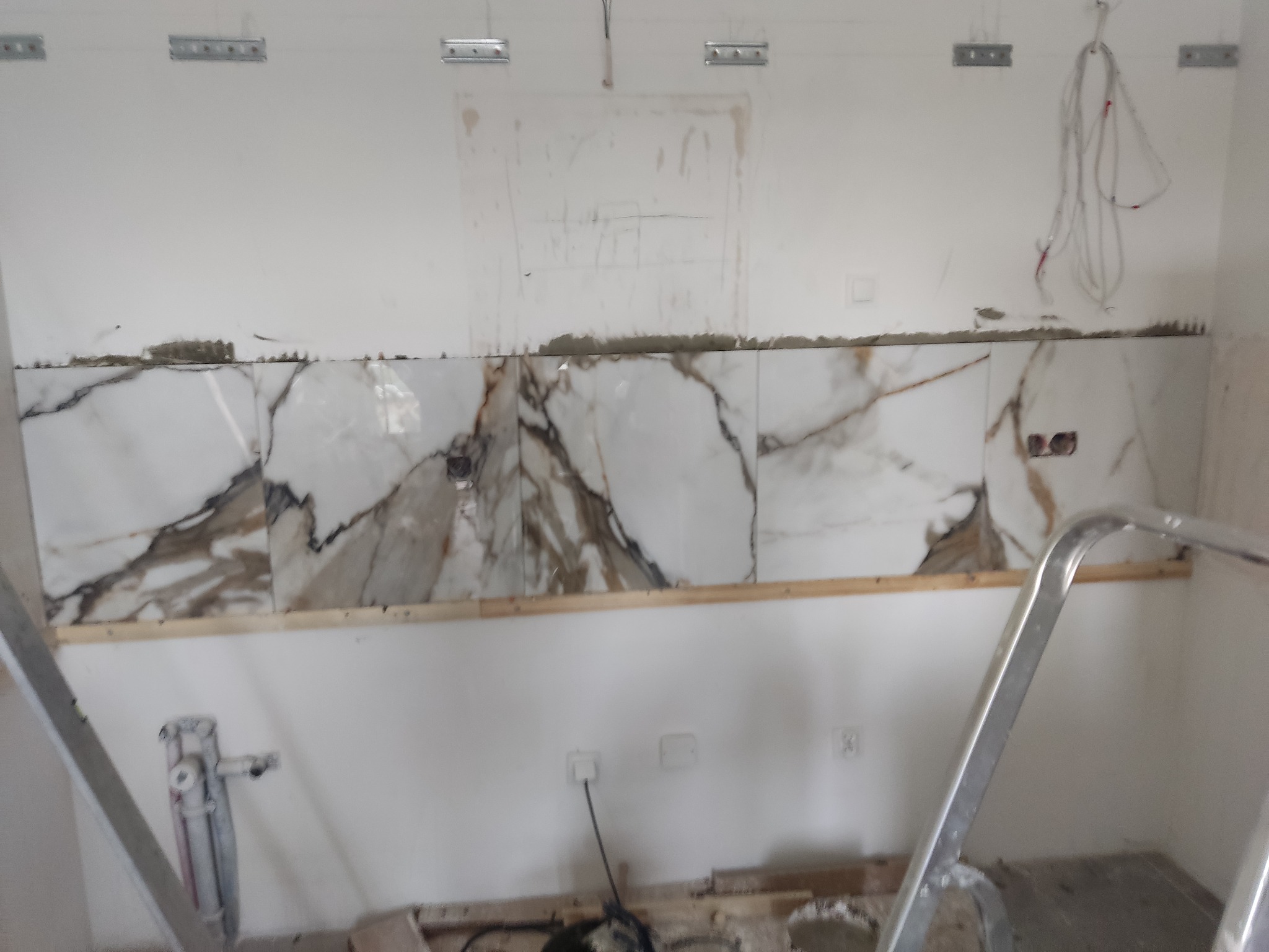 Ściana w trakcie montażu płytek imitujących marmur, widoczne metalowe profile sufitowe, przewody elektryczne i drabina malarska.