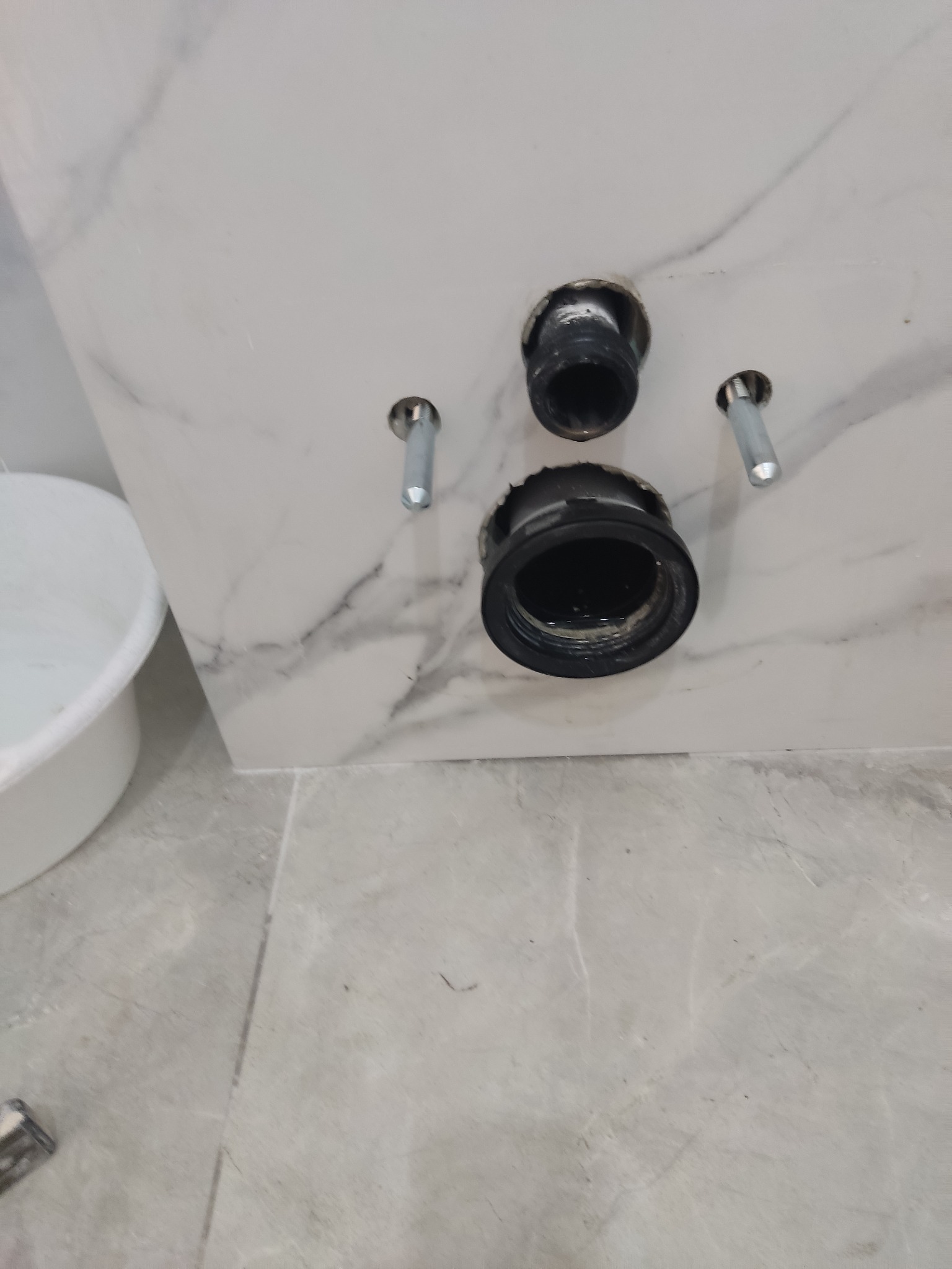 Przygotowane otwory kanalizacyjne i mocowania w marmurowej ścianie, widoczny fragment białego wiadra i szarej podłogi z płytek.