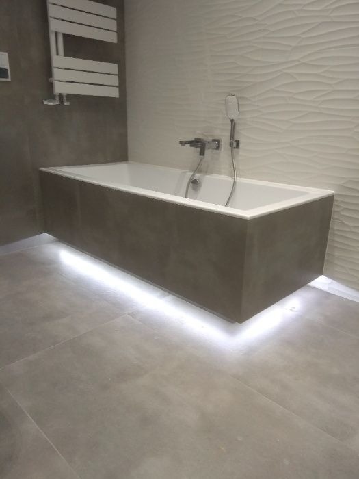 Nowoczesna łazienka z podświetlaną wanną w zabudowie, białe płytki o falistej strukturze na ścianie, szary gres na podłodze.