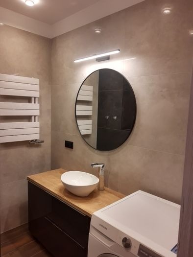 Nowoczesna łazienka z okrągłym lustrem podświetlonym listwą LED, umywalką nablatową na szafce z drewnianym blatem i białym grzejnikiem drabinkowym.