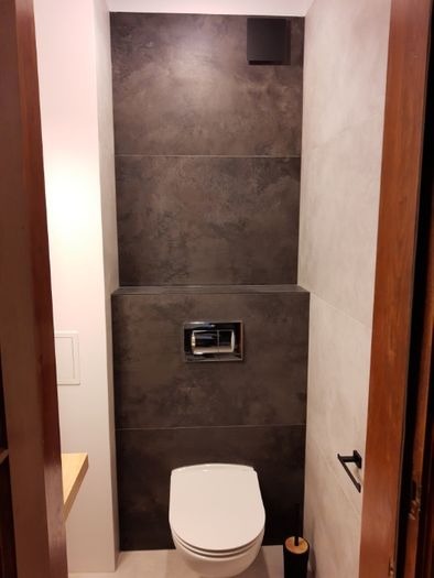 Nowoczesna toaleta z ciemnymi, strukturalnymi płytkami i chromowanym przyciskiem spłukującym, uzupełniona o białą miskę ustępową i czarne akcesoria.