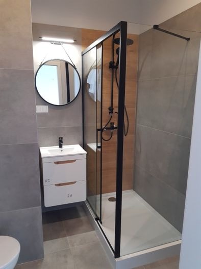 Nowoczesna łazienka po remoncie z kabiną prysznicową typu walk-in z czarnymi profilami, okrągłym lustrem z podświetleniem LED i białą szafką pod umywalką z szufladami.