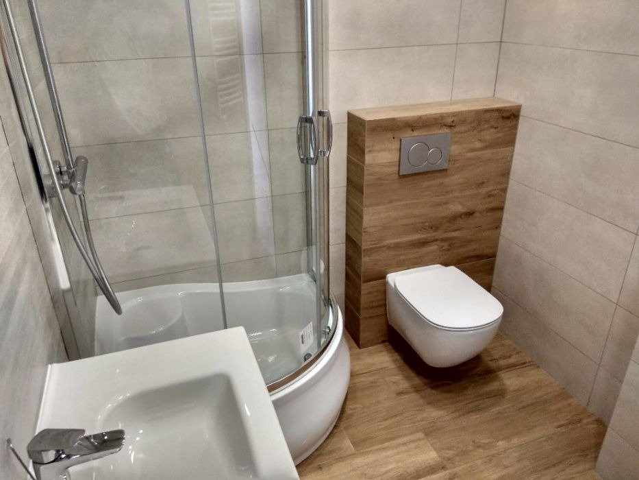 Nowoczesna łazienka po remoncie w Łomży: kabina prysznicowa z wanną, umywalka i podwieszany sedes na tle drewnopodobnych płytek.