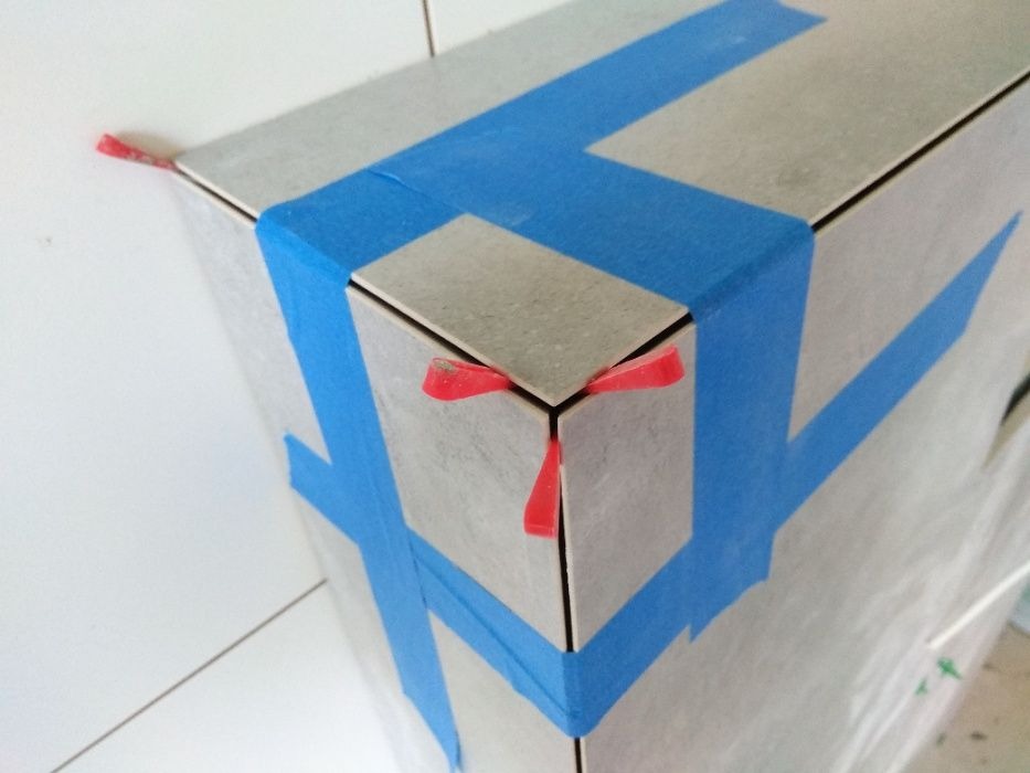 Fragment narożnika obłożonego płytkami, zabezpieczonego niebieską taśmą malarską i czerwonymi klinami dystansowymi.