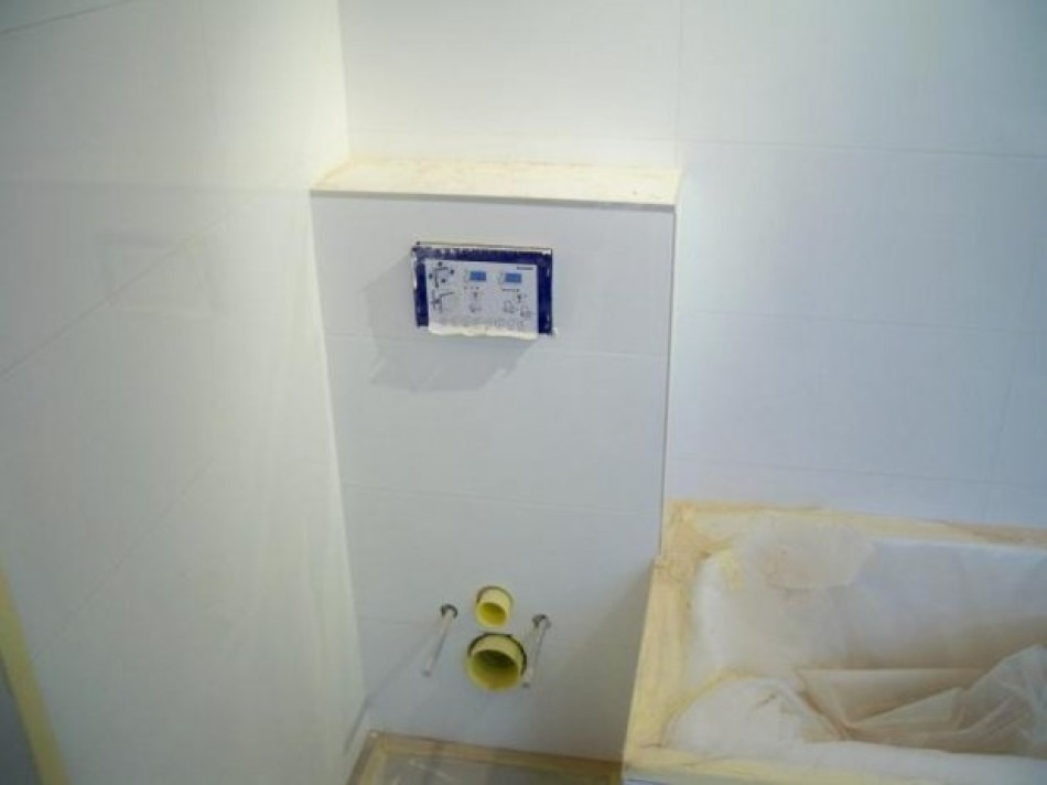 Białe płytki na ścianie w trakcie montażu stelaża podtynkowego WC, widoczne przyłącza kanalizacyjne i fragment zabezpieczonej folią wanny.