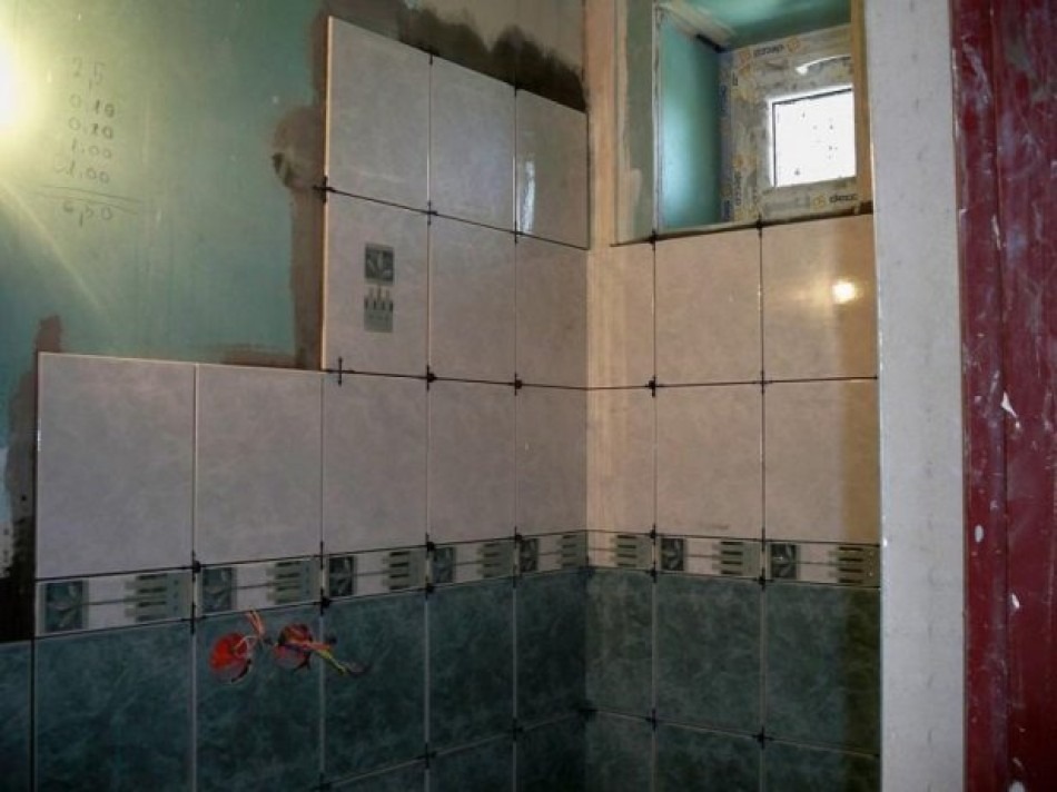 Układanie beżowych i zielonych płytek w łazience, widoczne krzyżyki dystansowe i niezakończone prace przy ścianie z oknem.