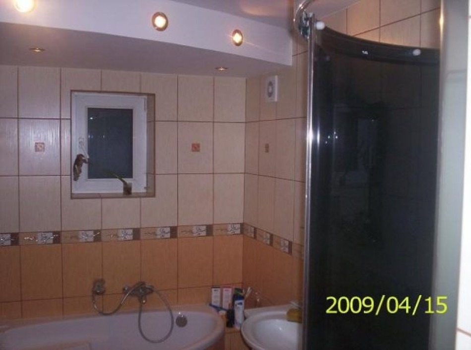 Łazienka z beżowymi i brązowymi płytkami, wanną, umywalką i kabiną prysznicową z ciemnego szkła; na ścianie okno z rośliną w doniczce.