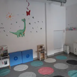 Pokój zabaw dla dzieci z naklejkami dinozaurów na ścianie, zabawkami kuchennymi i kolorowym dywanem w kropki.