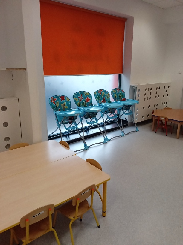 Jadalnia w placówce opiekuńczej: Dwa połączone stoliki z żółtymi nogami i dopasowanymi krzesełkami dziecięcymi z etykietami na oparciach, ustawione w pobliżu okna z pomarańczową roletą i rzędem...