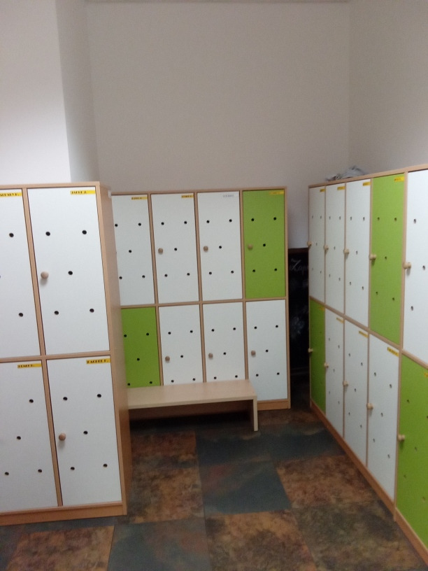 Szatnia z rzędami białych i zielonych szafek z drewnianymi uchwytami, uzupełniona o niską, drewnianą ławeczkę.
