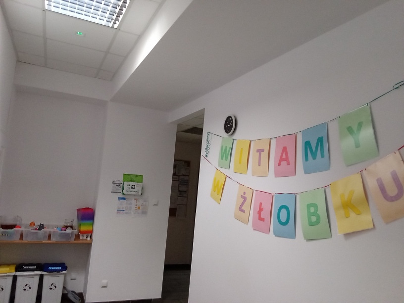 Wnętrze: ściana z dekoracyjnym napisem 'Witamy w ...', widoczne pojemniki na zabawki i apteczka pierwszej pomocy.