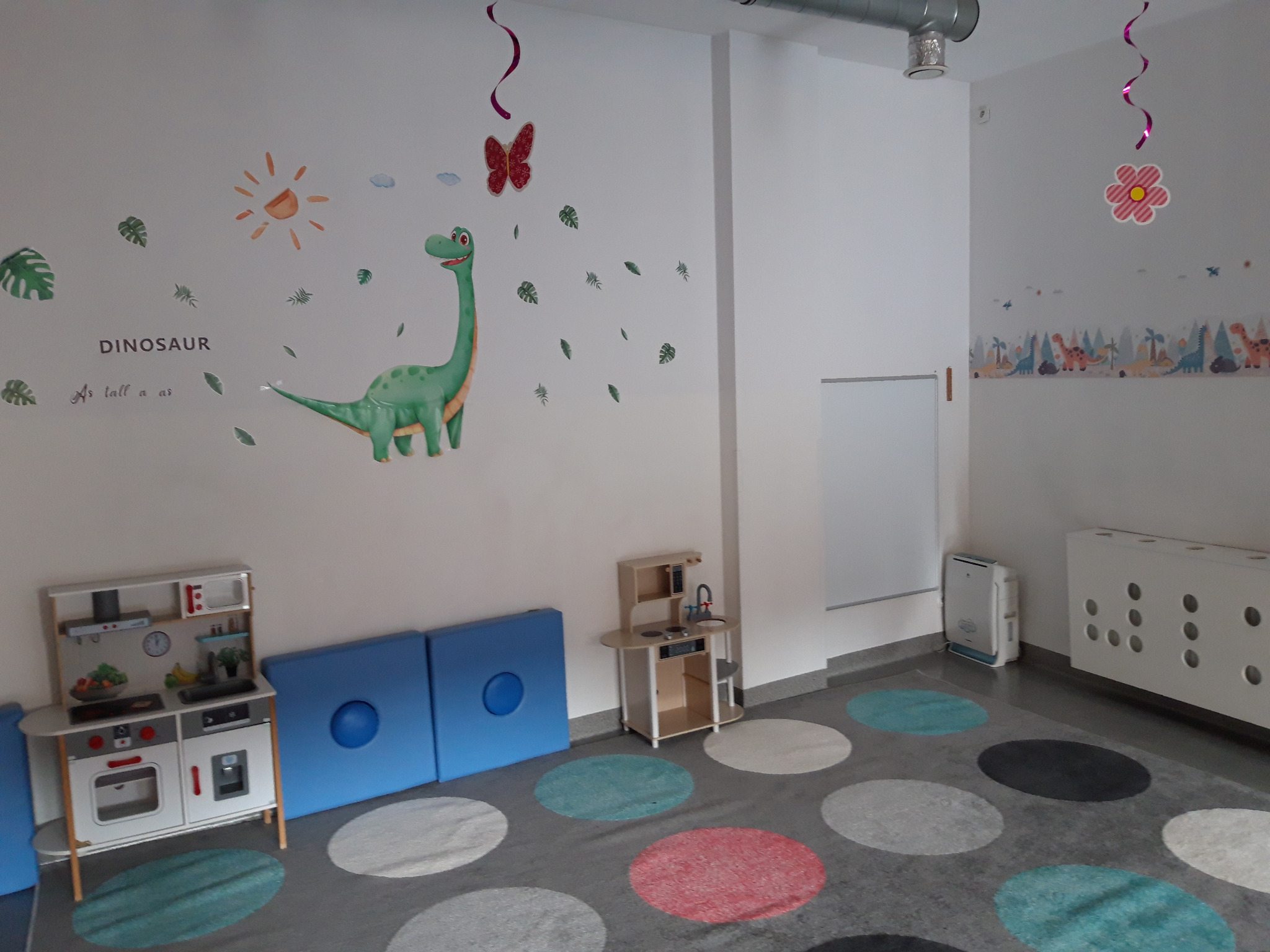 Pokój zabaw dla dzieci z naklejkami dinozaurów na ścianie, zabawkami kuchennymi i kolorowym dywanem w kropki.