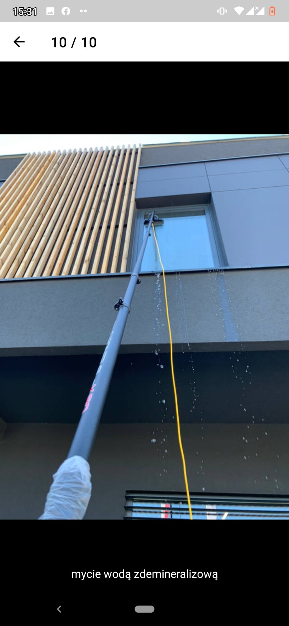 Mycie okna na wysokości za pomocą lancy teleskopowej z podawaniem wody demineralizowanej, widoczny budynek z drewnianymi elementami dekoracyjnymi.