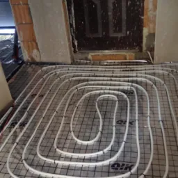 Białe rury ogrzewania podłogowego ułożone spiralnie na czarnej siatce w pomieszczeniu w trakcie remontu, widoczna surowa cegła i stara, ciemna, zabrudzona farbą stolarka drzwiowa.