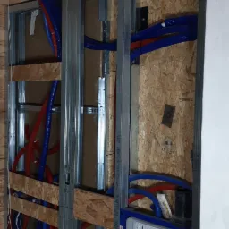 Stelaż podtynkowy WC marki Mexen, zabudowany w konstrukcji z profili stalowych, z widocznymi instalacjami hydraulicznymi w peszlach czerwonych i niebieskich, na tle ściany z płyty OSB i drewnianego...