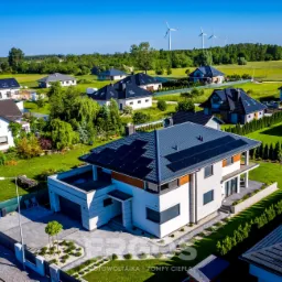 Widok z lotu ptaka na nowoczesny dom z panelami słonecznymi na dachu, na tle innych domów i turbin wiatrowych na horyzoncie.