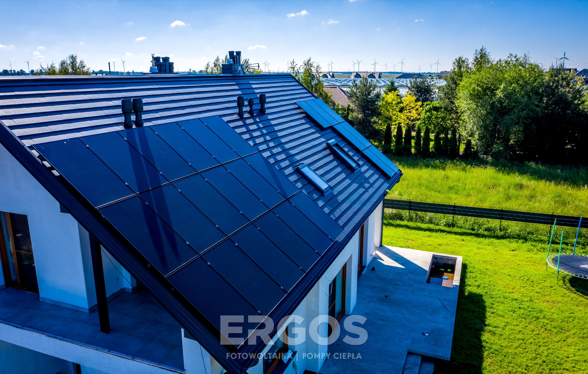 Czarny dach skośny z panelami słonecznymi i oknami dachowymi, na tle krajobrazu z farmą wiatrową w oddali.