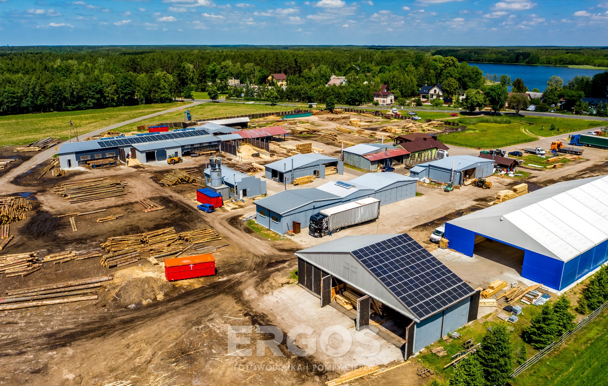 Widok z drona na zakład przetwórstwa drewna z zainstalowanymi panelami słonecznymi na dachach budynków, w tle las i jezioro.