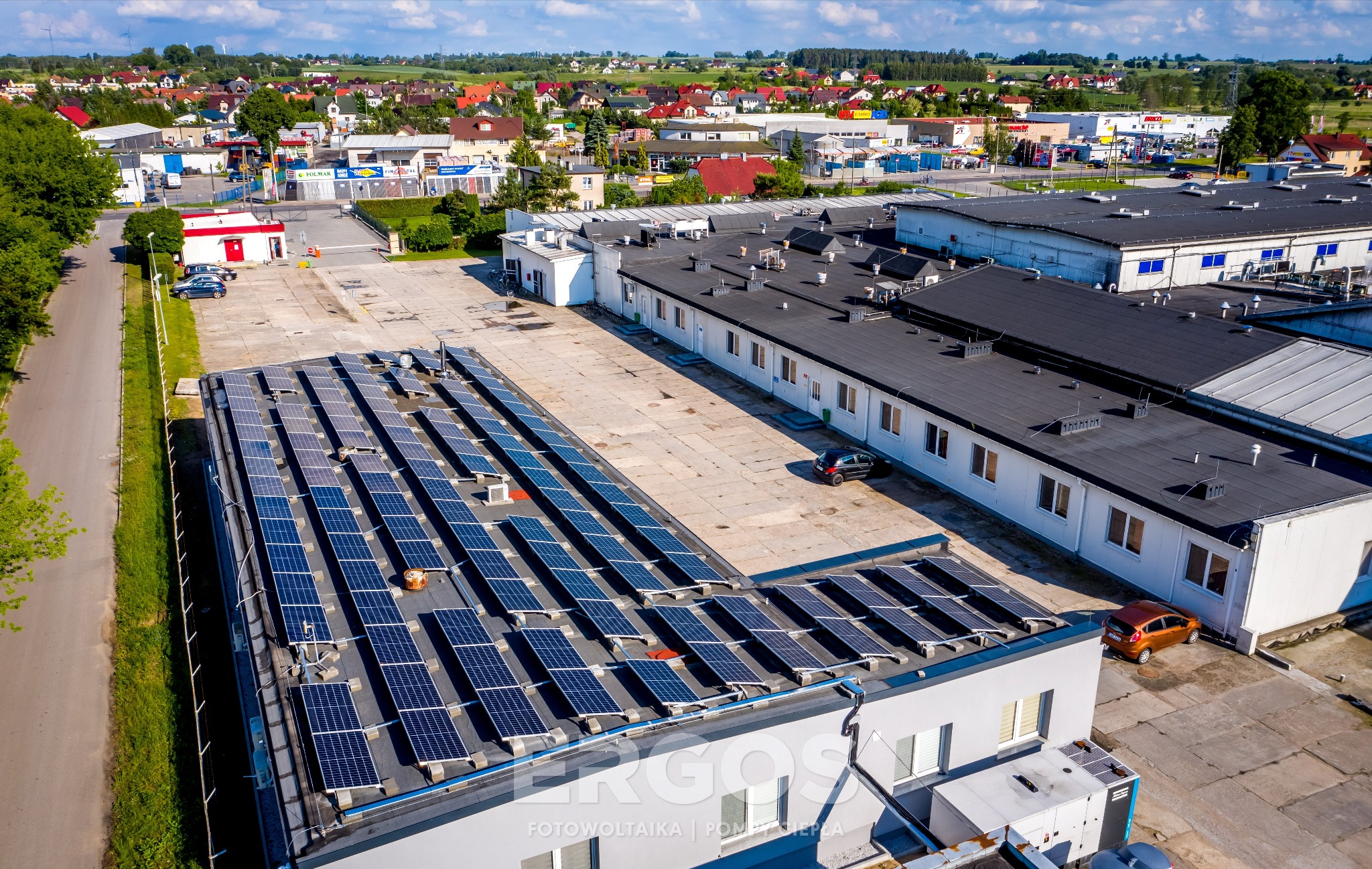 Widok z góry na budynek z panelami słonecznymi na dachu, w tle zabudowa miejska i drzewa, słoneczny dzień.