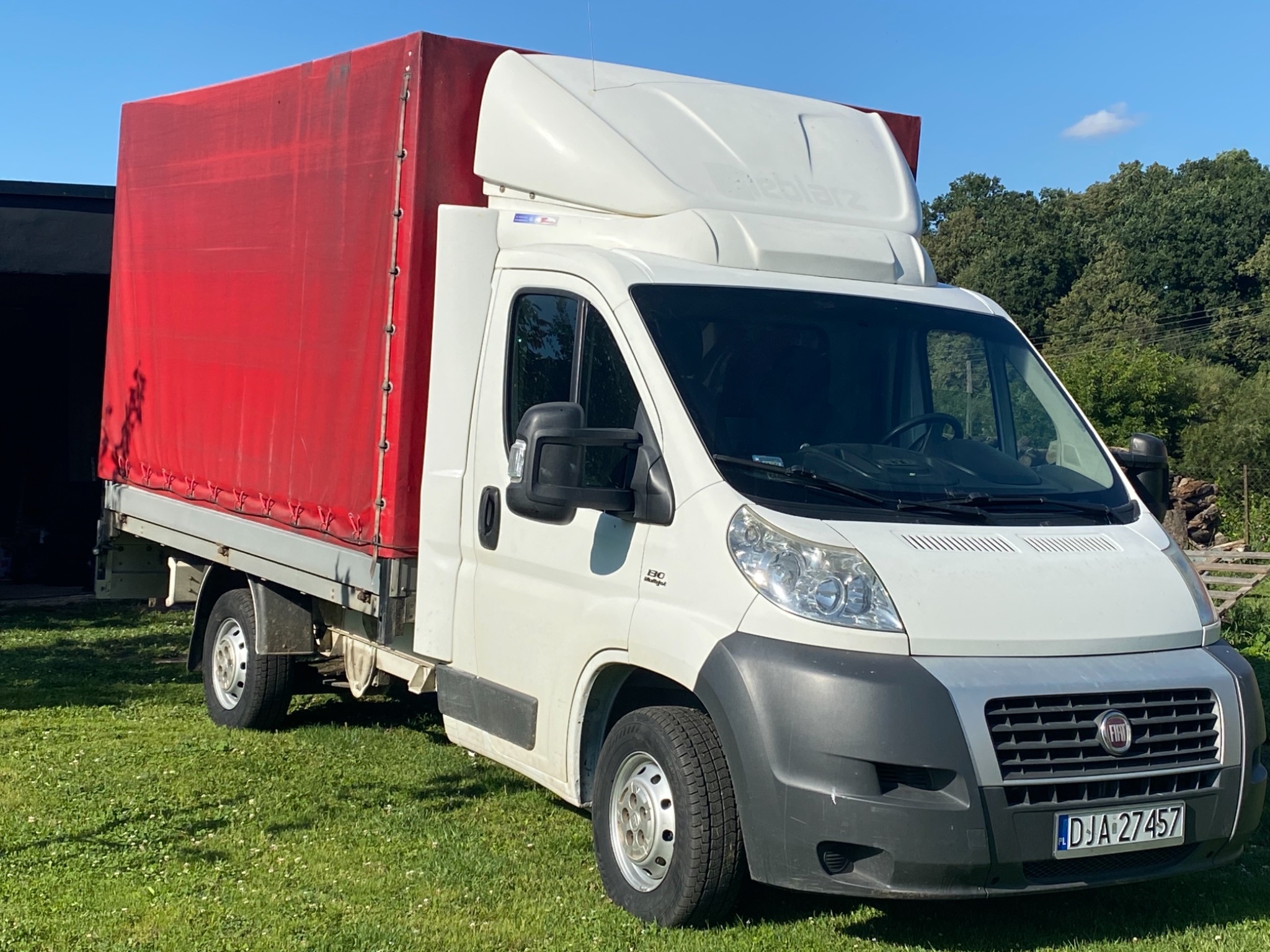 Biały samochód dostawczy z czerwoną plandeką na trawiastym terenie, widoczne tablice rejestracyjne i logo producenta.