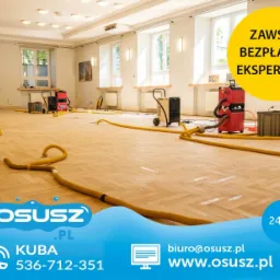 Parkiet w trakcie osuszania w Katowicach, widoczne żółte węże i profesjonalne osuszacze powietrza w dużym pomieszczeniu z oknami.