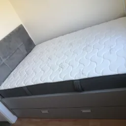 Szare łóżko z tapicerowanym zagłówkiem i białym materacem ze wzorem, wbudowane w szafę z szufladami.