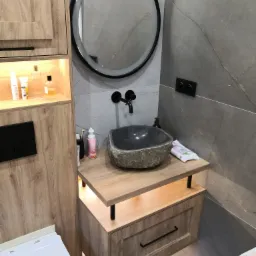 Nowoczesna łazienka z kamienną umywalką i podświetlaną szafką z drewnopodobnym wykończeniem, okrągłe lustro z podświetleniem LED i szare płytki imitujące marmur na ścianie.