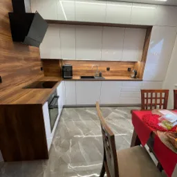 Nowoczesna kuchnia z białymi szafkami i drewnianym blatem, czarny okap nad płytą indukcyjną, oświetlenie LED podszafkowe i sufitowe, marmurowa podłoga, stół z czerwonym obrusem w tle.