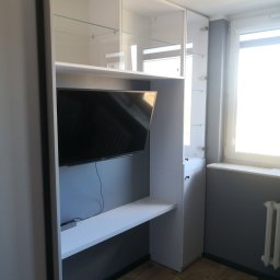 Eldar Meble Tomasz Szymborski - Biała szafa wnękowa z szklanymi półkami i miejscem na telewizor, uzupełniona blatem biurka. Nowoczesny design, drewniana podłoga, szara ściana.