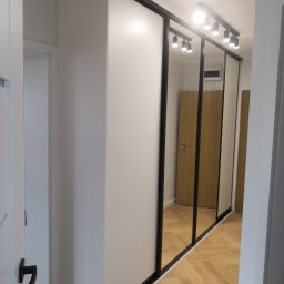 Eldar Meble Tomasz Szymborski - Biała szafa wnękowa z przesuwnymi drzwiami z czarnymi ramami i lustrami, oświetlona punktowo, na podłodze parkiet w jodełkę. Widok z perspektywy korytarza.