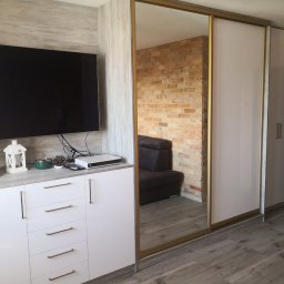 Eldar Meble Tomasz Szymborski - Biała szafa wnękowa z lustrzanymi drzwiami przesuwnymi, zintegrowana z szafką RTV. Widoczny fragment ceglanej ściany w odbiciu lustra.