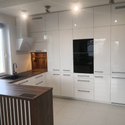 Eldar Meble Tomasz Szymborski - Biała, nowoczesna kuchnia z drewnianym blatem i wyspą. Fronty szafek na wysoki połysk, czarne, minimalistyczne uchwyty. Widoczny okap i zabudowany sprzęt AGD.